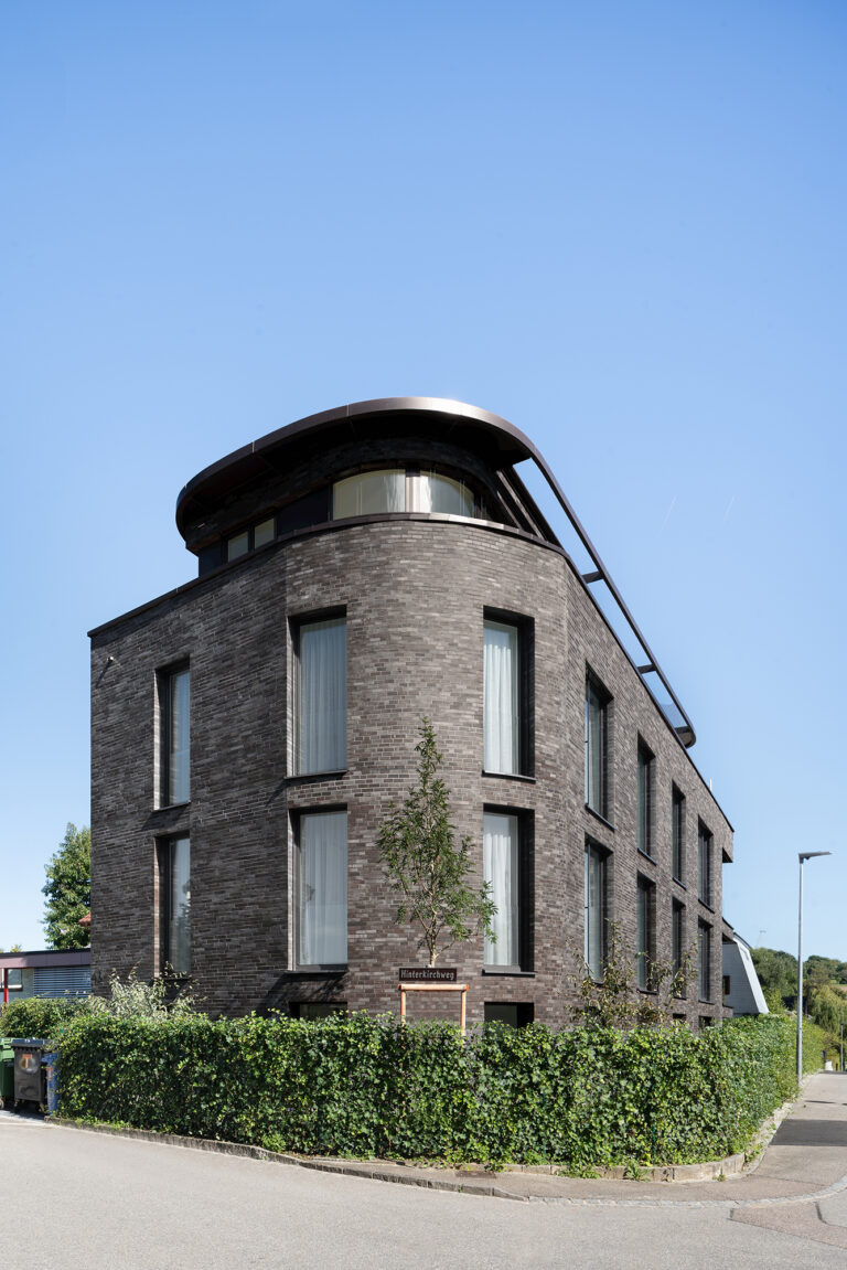 Foto von der abgerundeten Vorderansicht eines Mehrfamilienhauses mit dunklem warmen Klinker vor blauem Himmel.