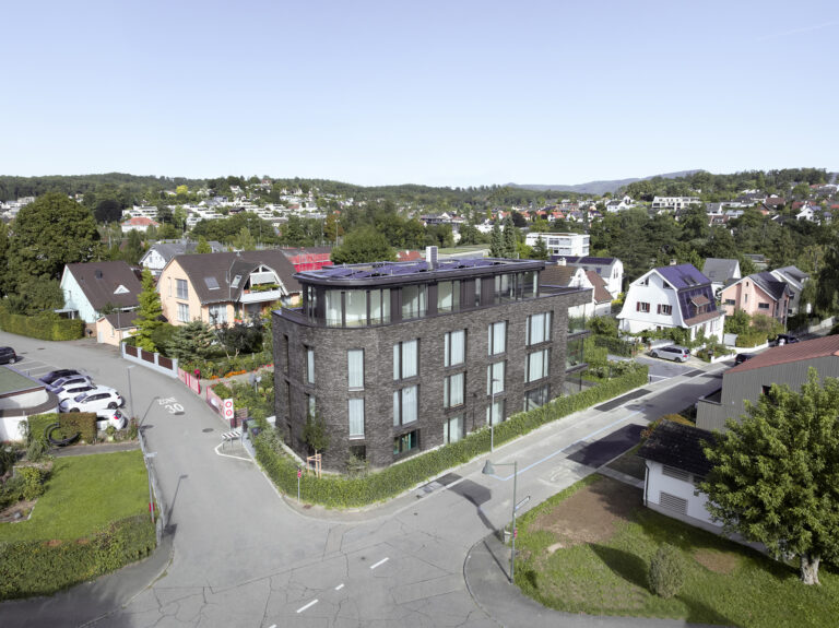 Foto einer Vogelperspektive eines vorne abgerundeten Mehrfamilienhauses mit nordischer Architektur und dunklem Klinker mit grossen Fensterfronten.