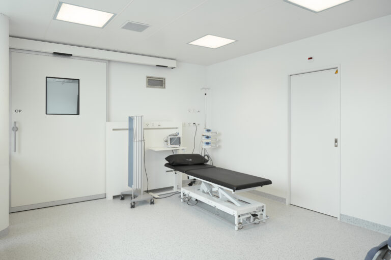 Foto eines Operationsraumes des Handzentrum mit weissen Wänden, Schiebetüre und schwarzer Liege.
