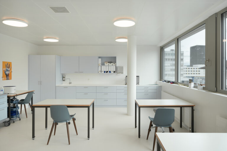 Foto eines hellerleuchteten Praxisraum und Blick auf eine Praxisküche und auf zwei Behandlungstischen von hinten und ein seitlicher Bürotisch. Am rechten Rand ist eine grosse Fensterfront zu sehen.