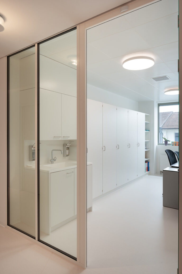 Foto mit Blick durch eine Glasfront in einen Behandlungsruam mit grossen weissen Schränken.