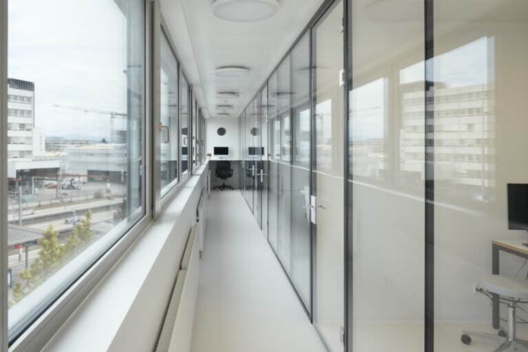 Foto eines Ganges zwischen langer Fensterfront und langer Glasfront mit Bürotisch am Ende des Ganges.