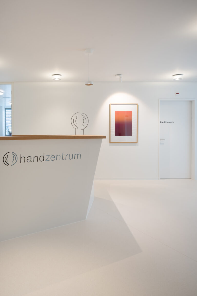 Ausschnitt-Foto einer hellen Praxis mit einer vorne abgeschrägter weisser Empfangstheke mit Holzablage und modernen Bildern, weissen Wänden und viel warmen Licht.