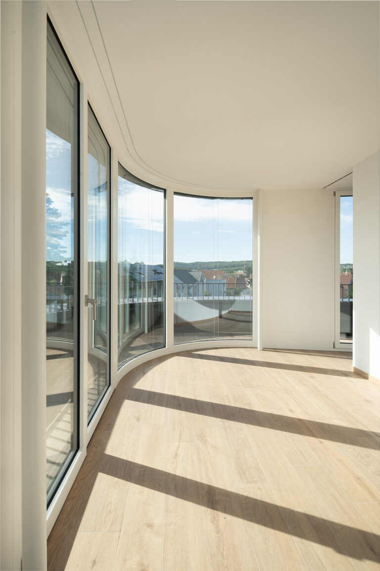 Foto eines lichtdurchfluteten warmen Raumes mit Parkettboden mit einer Fensterfront die im vorderen Teil abgerundet ist.
