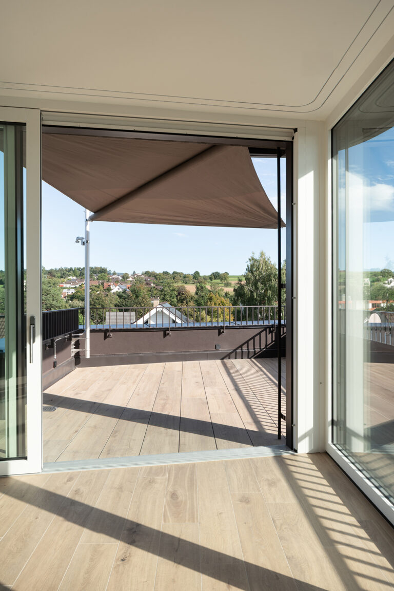 Foto-Ausschnitt eines Lichtdurchfluteten warmen Raumes mit Sicht auf die Terrasse und ausgezogener Markise und Blick ins Grüne.