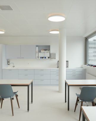 Foto eines hellerleuchteten Praxisraum und Blick auf eine Praxisküche und auf zwei Behandlungstischen von hinten und ein seitlicher Bürotisch. Am rechten Rand ist eine grosse Fensterfront zu sehen.