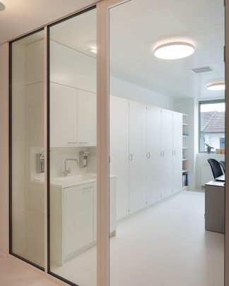 Foto mit Blick durch eine Glasfront in einen Behandlungsruam mit grossen weissen Schränken.