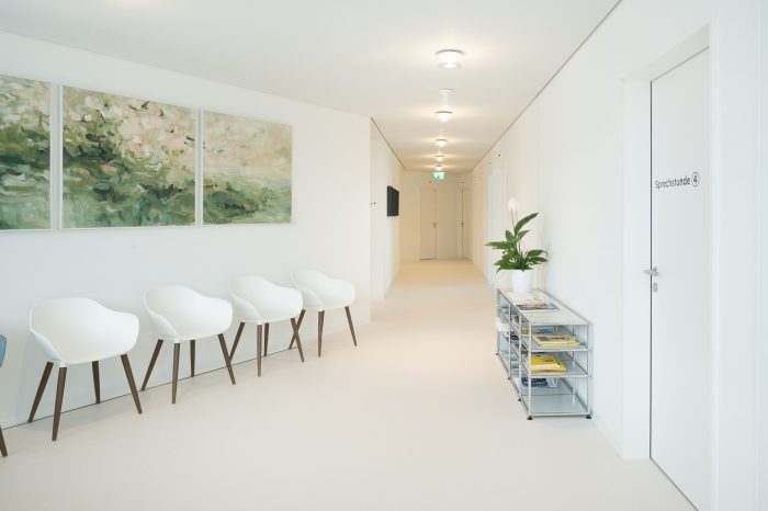 Foto eines hellen Warteraumes mit weissen Stühlen und ruhigen Bilder und einem hell und warm beleuchteten Ganges.