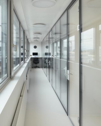 Foto eines Ganges zwischen langer Fensterfront und langer Glasfront mit Bürotisch am Ende des Ganges.