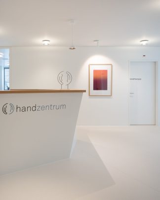 Ausschnitt-Foto einer hellen Praxis mit einer vorne abgeschrägter weisser Empfangstheke mit Holzablage und modernen Bildern, weissen Wänden und viel warmen Licht.