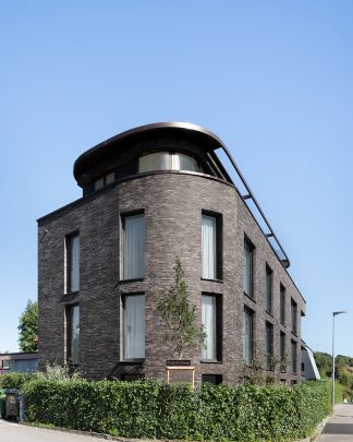 Foto von der abgerundeten Vorderansicht eines Mehrfamilienhauses mit dunklem warmen Klinker vor blauem Himmel.