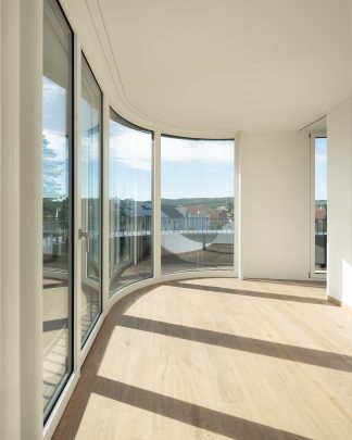 Foto eines lichtdurchfluteten warmen Raumes mit Parkettboden mit einer Fensterfront die im vorderen Teil abgerundet ist.