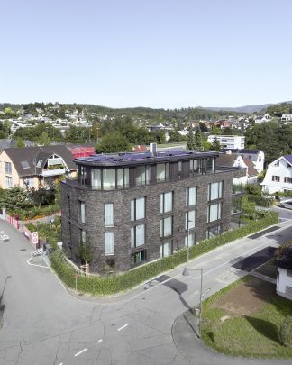 Foto einer Vogelperspektive eines vorne abgerundeten Mehrfamilienhauses mit nordischer Architektur und dunklem Klinker mit grossen Fensterfronten.