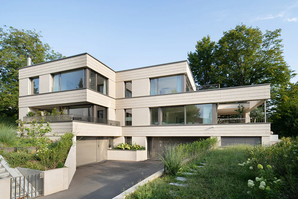 Frontansicht eines hellen, modernen Doppeleinfamilienhaus in Klinker mit grossen Fenster, vor grossen Bäumen.