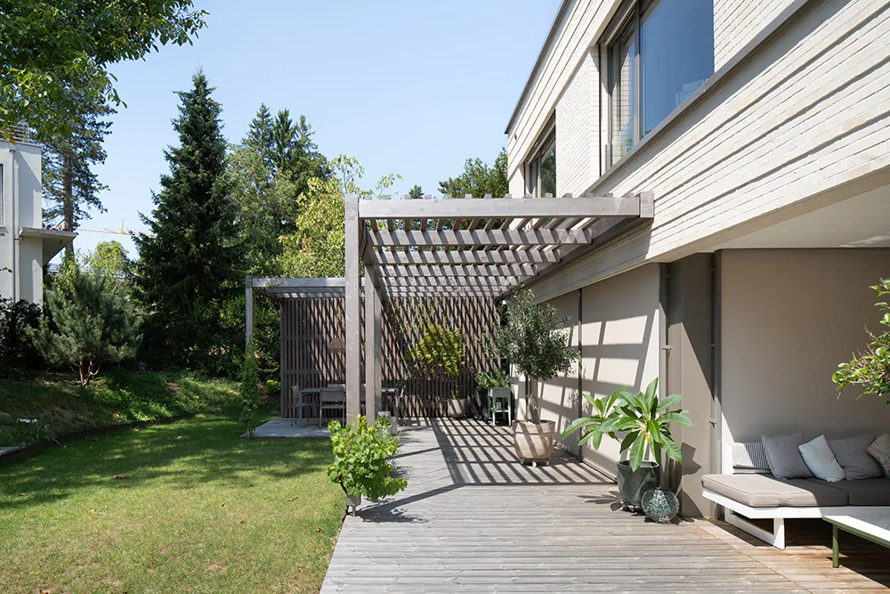 Gartenansicht eines hellen, modernen Doppeleinfamilienhaus in Klinker mit grossen Fenster mit Holzterrasse und Pergola in der Sonne.