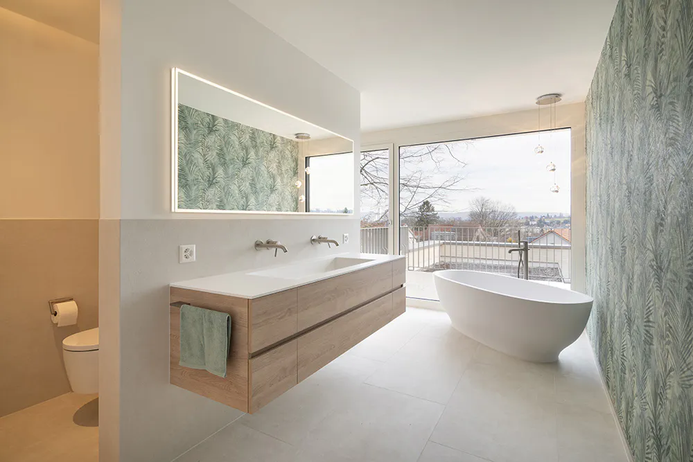 Sicht auf modernes offenes WC und Bad mit grossem Waschbecken in Holz und Spiegel mit grüner Tapette und grossem bodentiefes Fenster.